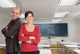 Ein Lehrer und eine Lehrerin stehen im Schulzimmer und lächeln in die Kamera.