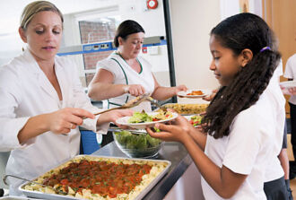 Zwei Köchinnen schöpfen einem Schulkind das Mittagessen auf einen Teller.