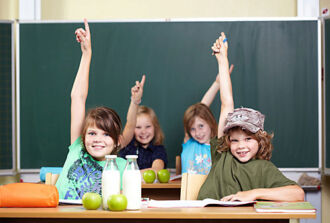 Vier Schulkinder sitzen an ihren Schulbänken. Alle strecken begeistert ihre Hand hoch, weil sie die Antwort auf eine vom Lehrer gestellte Frage wissen.
