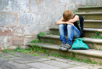 Ein Kind sitzt auf einer Treppe im Freien und hat seinen Kopf betrübt in seine auf den Knien aufgelegten Arme gesenkt.