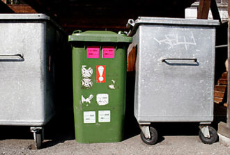 Ein Grüncontainer ist zwischen zwei blechernen Abfallcontainer eingeklemmt.